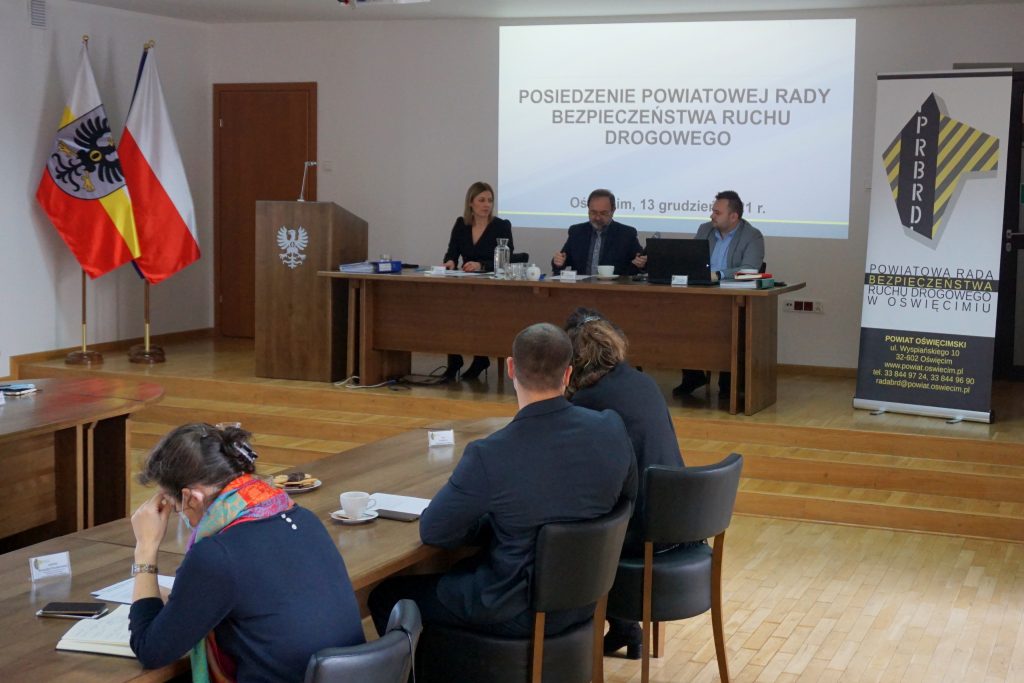 Sala konferencyjna trzy osoby siedzą pośrodku przy biurku. Widać stół przy którym siedzą osoby.