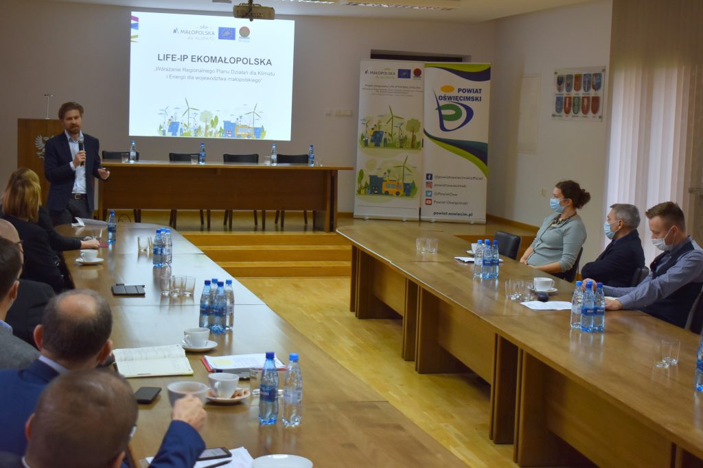 Sala konferencyjna po prawej i po lewej stronie stół przy nim siedzi kilka osób. Pośrodku ekran z prezentacją. Jedna osoba stoi i tłumaczy.