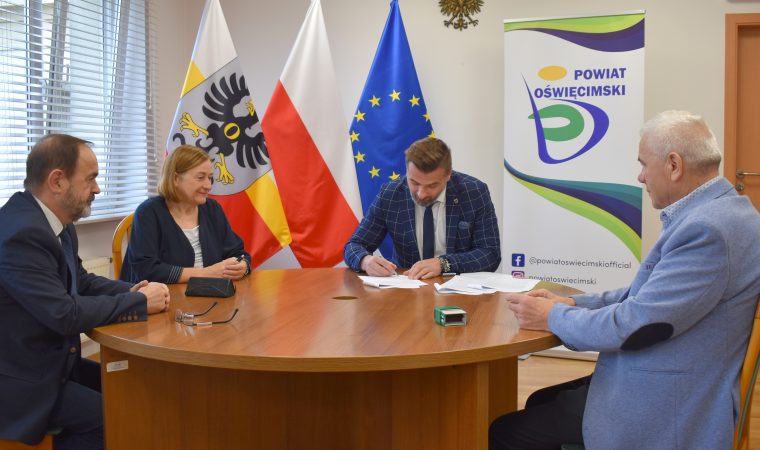 Cztery osoby elegancko ubrane siedzą przy okrągłym stole za nimi trzy flagi: Polski, Unii Europejskiej i Powiatu Oświęcim. Jedna z tych osób podpisuje dokumenty