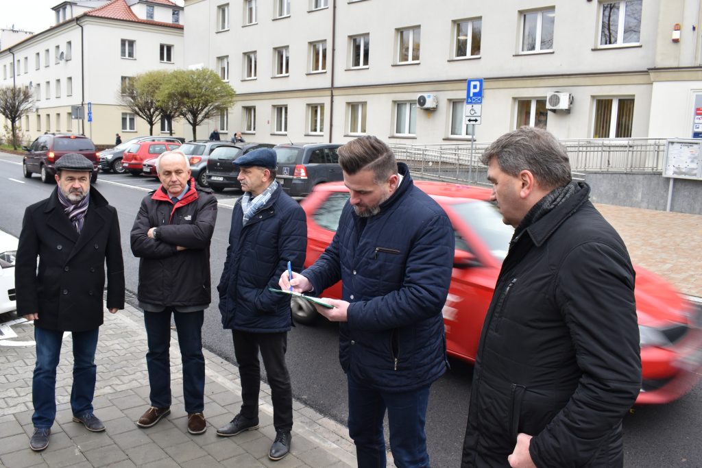 Pięć osób stoi, jedna z nich coś pisze na kartce, a za nimi przejeżdża czerwone auto.
