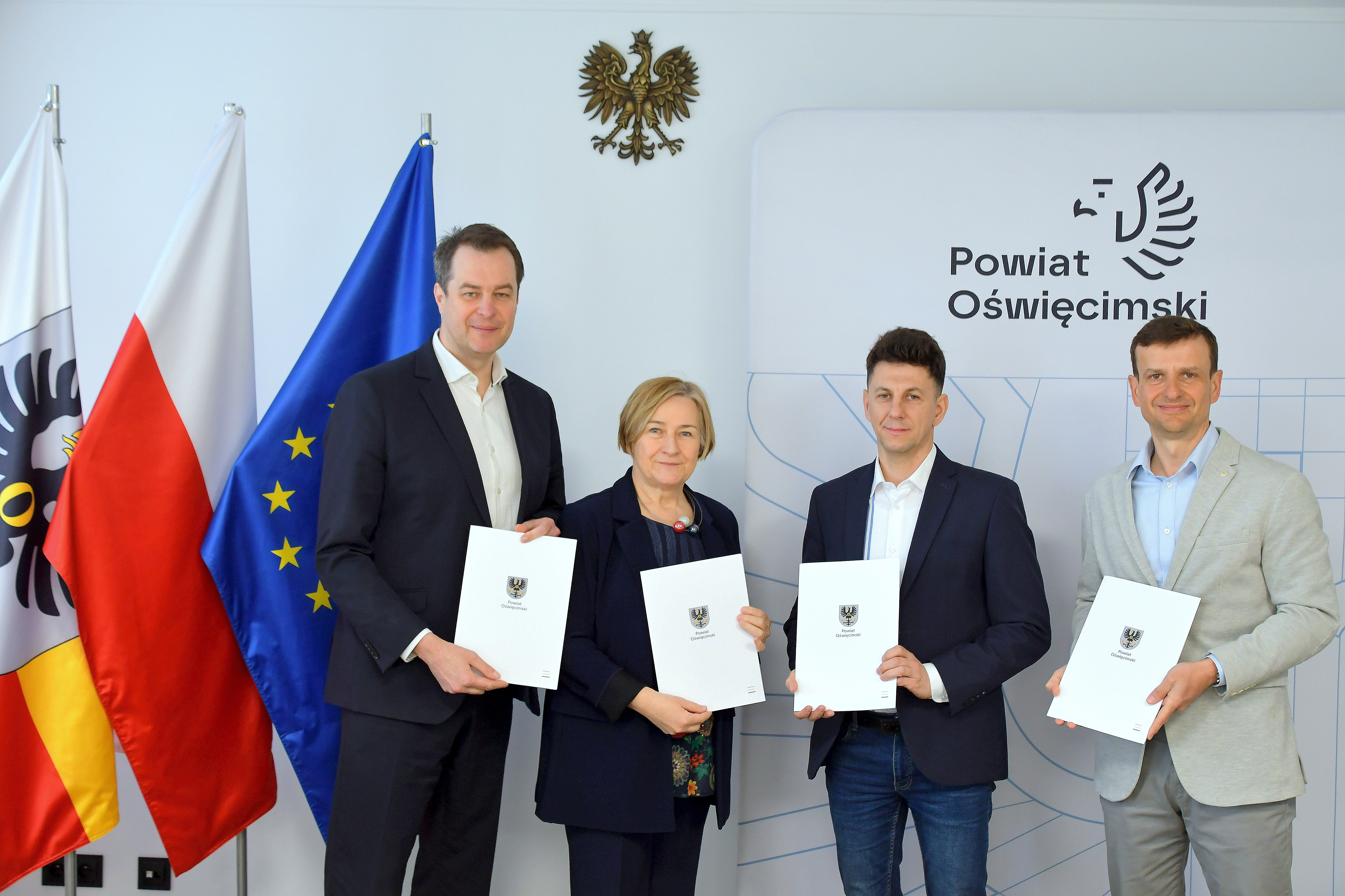 Pamiątkowe zdjęcie sygnatariuszy umowy na tle flag i logo Powiatu