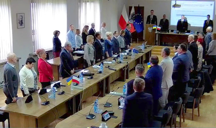 Radni Rady Powiatu w Oświęcimiu stoją w sali obrad i oddają hołd ukraińskim ofiarom rosyjskiego okrucieństwa.