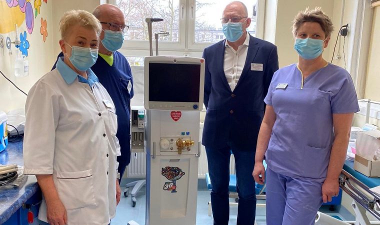 Zdjęcie, pośrodku nowe urządzenie szpitalne. Dookoła niego osoby trzy ubrane w stroje lekarskie, jedna w garniturze.