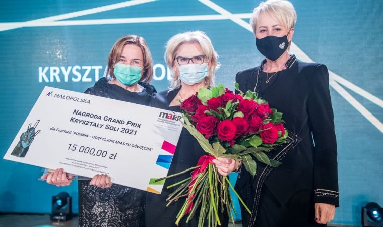 Pośrodku trzy osoby. Jedna osoba w środku trzyma kwiaty i nagrodę.