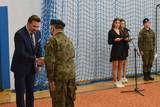 Dwie osoby jedna w mundurze druga w garniturze podają sobie dłonie. Obok po prawej stronie widać osobę przemawiająca do mikrofonu