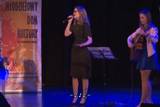 Pośrodku osoba śpiewa na scenie po prawej stronie druga osoba gra na gitarze.