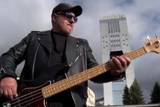 Mężczyzna grający na gitarze elektrycznej. W tle wysoki budynek.